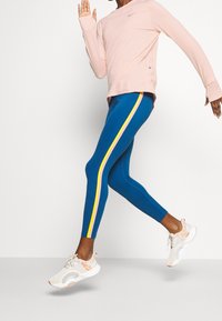 Blaue Leggings mit gelben und rosa Seitenstreifen, kombiniert mit einem hellrosa Langarmoberteil und hellen Sneakers. Glattes, dehnbares Material.