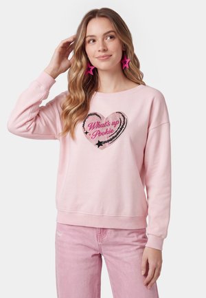 Ung kvinde iført en pink sweatshirt med hjerte og "What's up Pookie" tekst, matchende pink stjerne øreringe og lyserøde bukser.