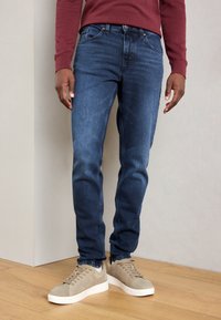 Skinny-fit denim jeans in dark blue with a slight fade, featuring a classic five-pocket design and contrast stitching. Paired with light gray sneakers.