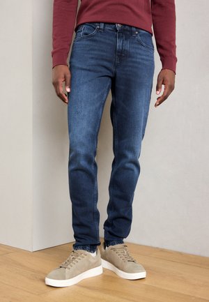 Skinny-fit denim jeans in donkerblauw met een lichte vervaging, voorzien van een klassieke vijf-pocket ontwerp en contrasterende stiksels. Gecombineerd met lichtgrijze sneakers.