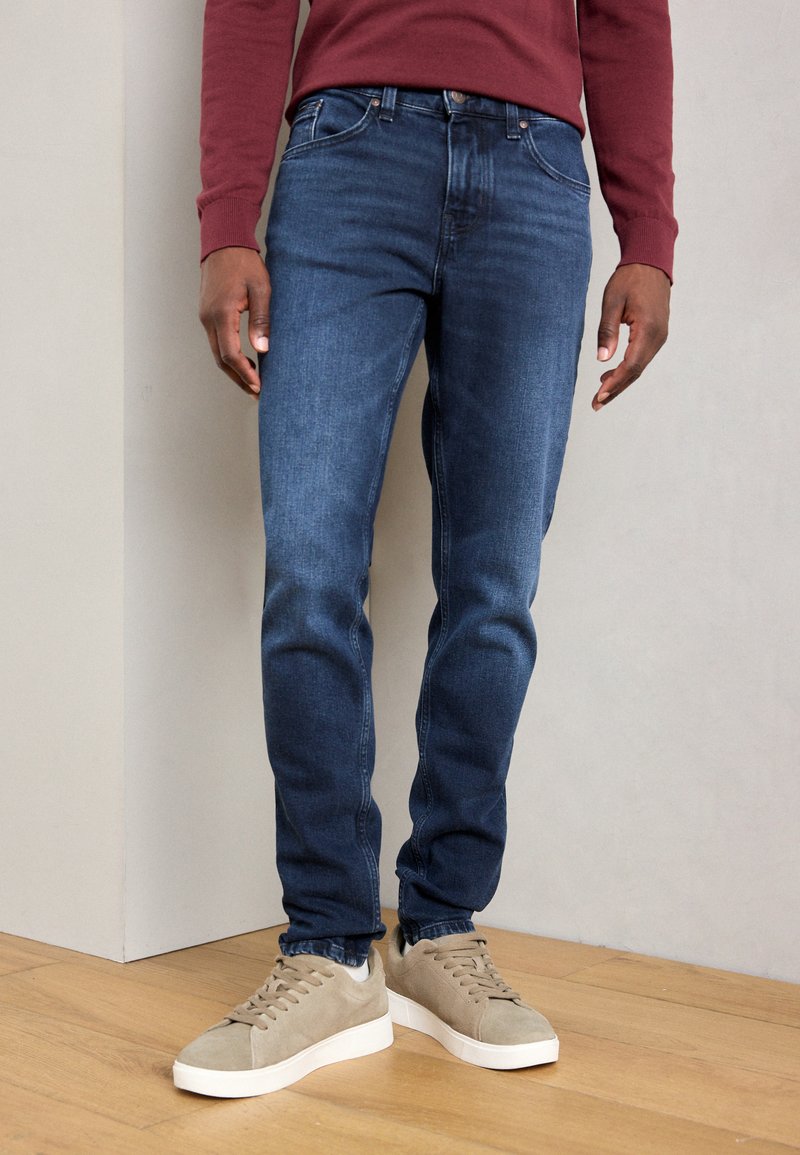 Skinny-fit denim jeans in dark blue with a slight fade, featuring a classic five-pocket design and contrast stitching. Paired with light gray sneakers.
