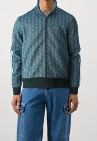 Grön zip-up-jacka med geometriskt mönster, ribbade ärmslut och blå sidostrippar. Har Lacoste-logotyp på bröstet.