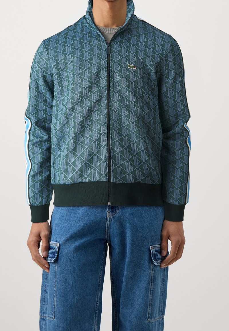 Grön zip-up-jacka med geometriskt mönster, ribbade ärmslut och blå sidostrippar. Har Lacoste-logotyp på bröstet.