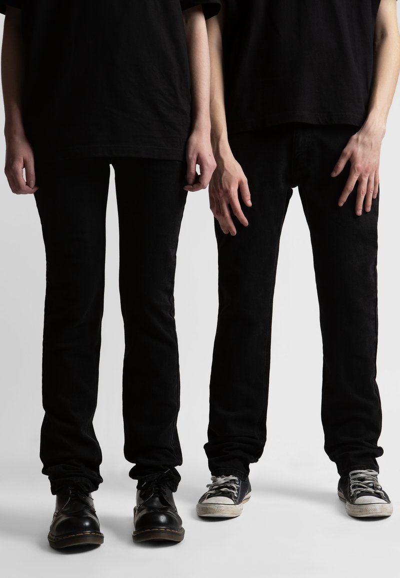 Δύο όρθιοι άνθρωποι που φορούν μαύρο παντελόνι και μαύρα πουκάμισα, ο ένας με παπούτσια φόρμας και ο άλλος με φθαρμένα αθλητικά παπούτσια, εμφανιζόμενοι από τον λαιμό μέχρι τα πόδια.