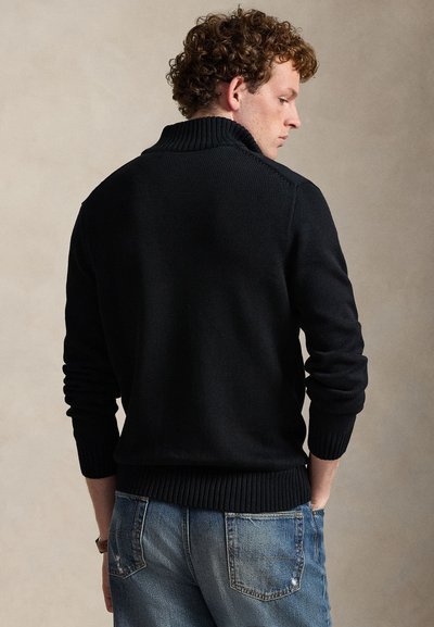 Mann mit lockigem Haar, der einen schwarzen gerippten Pullover und blaue Jeans trägt, steht mit dem Rücken zu einem einfarbigen beigen Hintergrund.