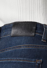 Tummansiniset farkut, joissa on musta nahkainen merkintä, johon on prässätty teksti "DKNY JEANS". Vyötäröllä näkyvät oranssit tikit.