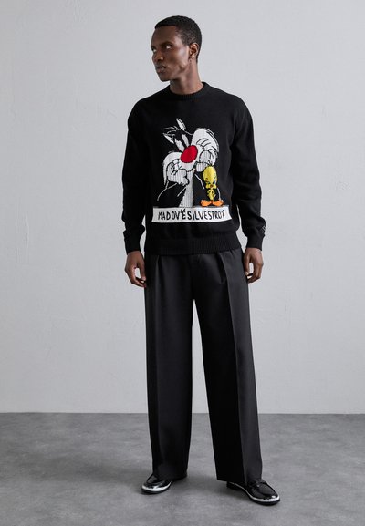 Pull noir en tricot avec un design de bande dessinée représentant Sylvester le Chat et Tweety Bird, associé à un pantalon noir ample et des chaussures brillantes.