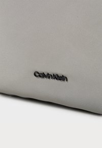 Siva teksturirana tkanina torbica s glatkom završnom obradom, sa crnim „Calvin Klein“ logotipom urezanim na površini.