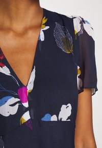 Marineblaues Blumenkleid mit kurzen, durchsichtigen Ärmeln; enthält weiße, rosa und gelbe Blumenmuster; V-Ausschnitt; strukturierter Stoff mit einem dezenten Glanz.