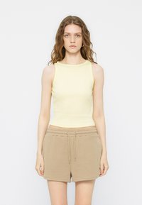 Γυναίκα με κυματιστά μαλλιά, που φορά αμάνικη ανοιχτό κίτρινη μπλούζα και μπεζ σορτς με κορδόνι, στέκεται μπροστά σε ένα απλό λευκό φόντο.