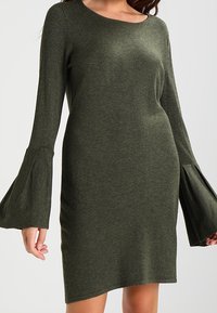 Olivgrön klänning i mjukt stickat tyg, med långa trumpetärmar och rund halsringning, med en figurnära silhuett och asymmetrisk nederkant.