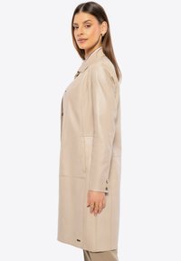 Beige leren jas met een op maat gemaakte snit, voorzien van een kraag, zijzakken en knoopdetails, gepresenteerd in een strakke, gladde textuur.
