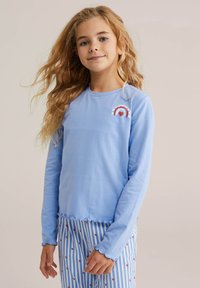 Camisa de manga larga azul claro con dobladillo y puños ondulados; presenta un diseño gráfico de arcoíris con un detalle de corazón en el pecho izquierdo.
