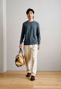 En mann har på seg en blå genser og kremfargede bukser, og holder en beige veske med mørke detaljer. Rommet har et lysekt tremgulv og nøytrale vegger.