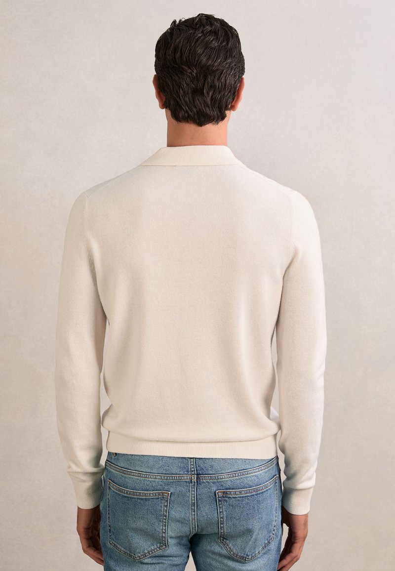 Homme aux cheveux courts et foncés portant un polo crème à manches longues et un jean bleu, debout, dos tourné, sur un fond uni.