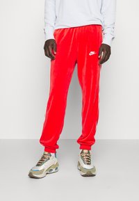 Červené sametové tepláky s elastickými manžetami a pasem. Obsahují bílé logo Nike a postranní pruhy. Na nohou má multicolor tenisky.