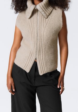 Gilet sans manches en maille côtelée beige avec col haut et fermeture éclair à l'avant, porté avec un pantalon noir ample.
