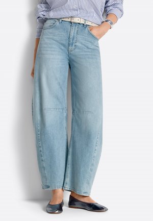 Donna con jeans a gamba larga azzurri, camicia a righe, cintura bianca, ballerine nere e braccialetti, con una mano in tasca.