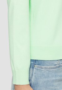 Jasnozielony sweter dzianinowy z prążkowanymi mankietami i dołem, w połączeniu z niebieskimi dżinsami, które mają mały metalowy akcent na kieszeni.