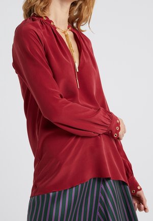 Kobieta ubrana w czerwoną bluzkę z długim rękawem z złotymi guzikami i złoty łańcuszek na szyi, w duecie z pasiastą zielono-fioletową spódnicą.