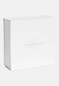 Bijela pravokutna kutija s glatkom, sjajnom površinom. Ugravirani logotip "Calvin Klein Jeans" u sivoj boji sprijeda. Kutija s preklopom na vrhu.