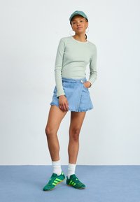 Langærmet lysegrøn top, blå denim omviklingsskirt, grønne sneakers med gule striber, hvide strømper. Model står på blå gulv mod en hvid væg.