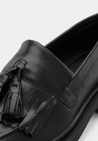 Musta nahkakengät tupsuakkenteilla, joissa on sileä pinta ja ommeltu yksityiskohta. Pohja on litteä ja hieman teksturoitu.