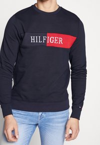 Námořnický svetr se červenobílým logem "HILFIGER" na hrudi. Vyroben z měkkého materiálu, má kulatý výstřih a žebrované manžety.