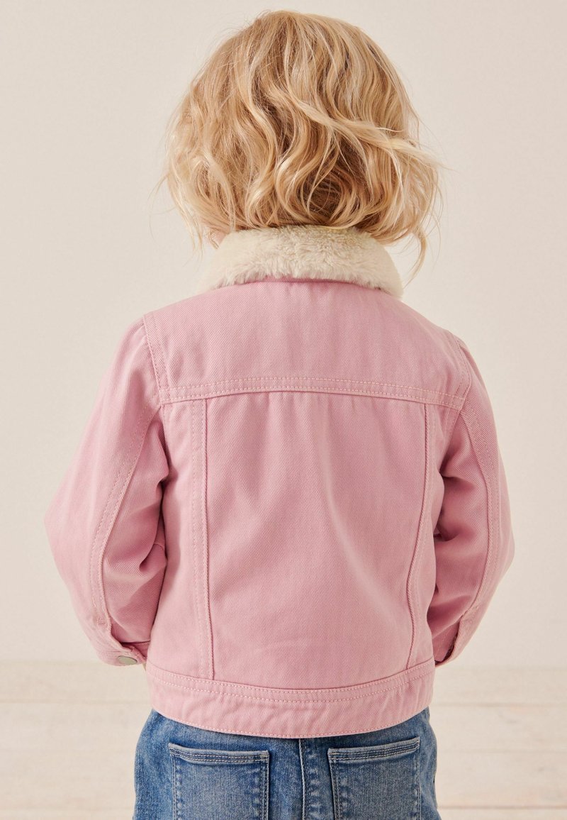 Enfant aux cheveux blonds ondulés portant une veste rose avec un col en fourrure blanche et un pantalon en jean bleu, dos à la caméra.