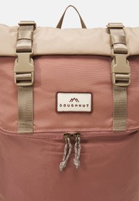 Doughnut backpack in soft pink and beige fabric with a roll-top design, gold hardware, and a front zip pocket with dual pulls.