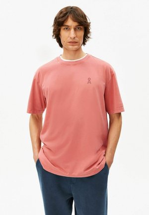 Man met halflang bruin haar, draagt een verweerd roze T-shirt en donkerblauwe broek, met handen in de zakken, tegen een effen witte achtergrond.