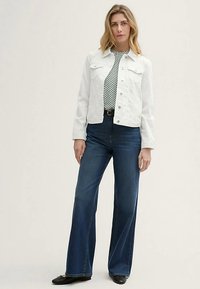 Witte spijkerjasje met knopen aan de voorkant en borstzakken, gedragen over een zwart-wit gestipte top. Wijde donkere spijkerbroek en zwarte platte schoenen.