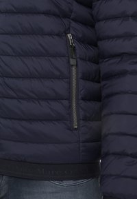 Marineblå pufferjakke med quiltet tekstur, udstyret med en lynlåslomme og en elastisk talje med branding. Glat, letvægtsstof.