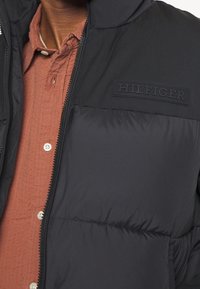 Homme portant une doudoune noire avec le logo « HILFIGER » sur la poitrine, par-dessus une chemise boutonnée couleur rouille.