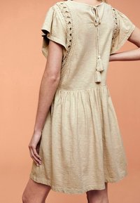 Beiges, kurzärmeliges Kleid mit Spitzenakzenten, gerafft an der Taille und dekorativen Quasten an der Rückseite. Weicher, strukturierter Stoff.