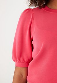 Rosa topp med puffärmar i mjukt tyg, med rund halsringning och rynkad detalj vid ärmarna.