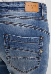 Denimjeans i ljusblått med en texturerad yta. Har en läderlogotyp, synlig sömnad och en bakficka med en metallknapp.