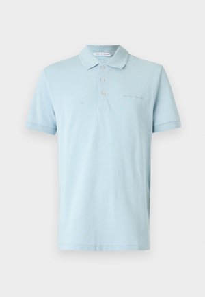 Man with short blonde hair wears light blue polo shirt, navy shorts, and brown sandals standing against plain white background.
