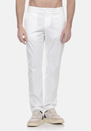 Hombre vistiendo pantalones blancos de pierna recta y zapatillas beige, de pie contra un fondo claro y liso.