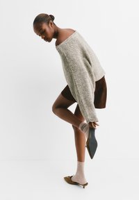Beiger Strickpullover, braune Wildleder-Rock und braune Absatzschuhe mit einer Schleife. Modell hält den Schuh, während sie ihre Socke anpasst. Schlichter Hintergrund.