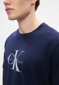 Jeune personne portant un T-shirt bleu marine à col rond avec le logo "Calvin Klein" bleu clair et blanc sur la poitrine.