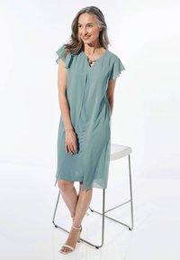 Ein fließendes türkisfarbenes Kleid mit kurzen Rüschensäumen, einem Schlitz vorne und einem leicht lockeren Schnitt, kombiniert mit weißen Absatzsandalen.