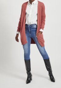 Rosa kofta med långa ärmar, tillsammans med en vit skjorta och blå jeans. Svarta klackar med krokodilprägling fulländar outfiten.