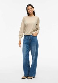 Beige longsleeve trui met geribde manchetten, gecombineerd met hoge taille wijde blauwe jeans. De outfit wordt aangevuld met puntige beige schoenen.