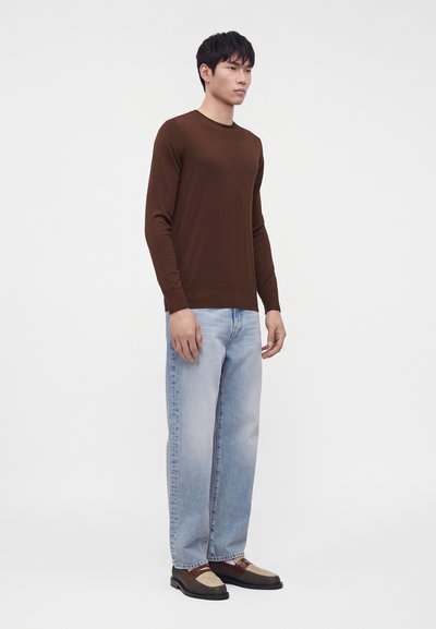 Homme portant une chemise marron à manches longues, un jean bleu clair et des chaussures marron et beige, debout de côté sur un fond blanc uni.