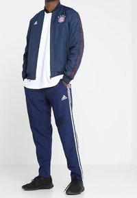 Navy bomber jacket with red accents and logo on chest, paired with navy track pants featuring white stripes along the sides. Black footwear.