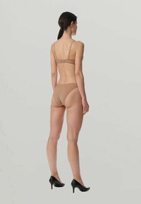 Femme aux cheveux bruns, en soutien-gorge et culotte nude, portant des talons hauts noirs, se tenant debout et dos à un fond gris uni.