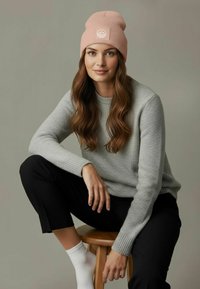 Light pink ribbed knit beanie with a white logo, paired with a gray sweater and black pants. Model seated on a wooden stool.