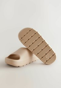 Sandale à glissière beige avec un bout ouvert et un design arrondi, fabriquée à partir d'un matériau texturé ; la semelle présente des motifs de grip circulaires et des trous de drainage.