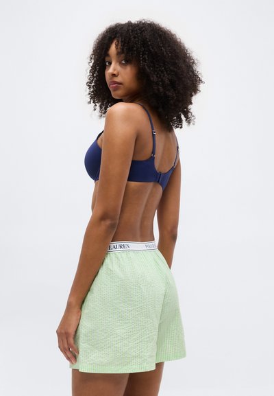 Γυναίκα με σγουρά μαλλιά, φορώντας μπλε σουτιέν και ανοιχτό πράσινο σορτς με υφή και ζωνάκι Polo Ralph Lauren, που στέκεται μπροστά σε λευκό φόντο.